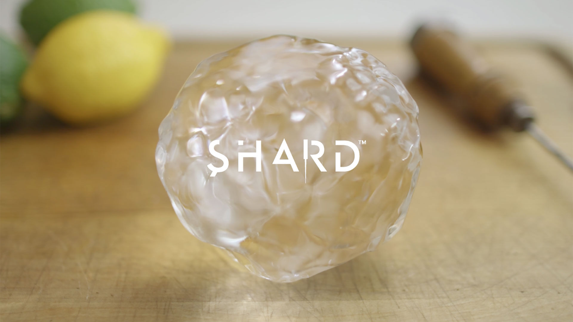 Hand-carved ice ball on a cutting board overlaid with the Shard logo, with lemon, lime, and ice pick in the background